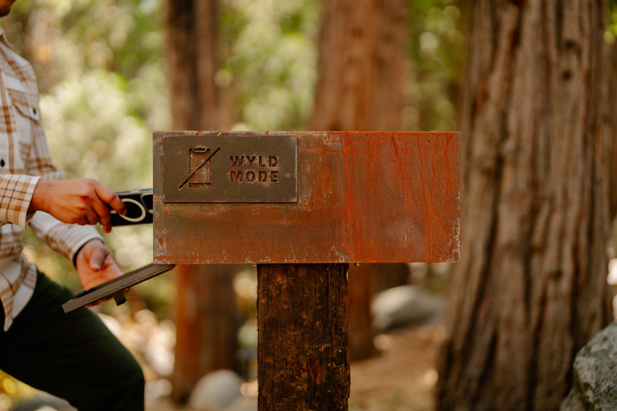 Guest placing phone in the rust-patina Wyld Mode box mounted on a wooden post among the Idyllwild pines
